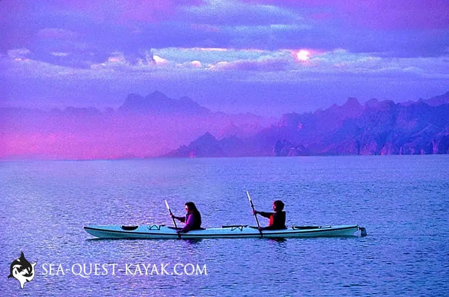 Two people kayaking on calm water with mountains and a purple sky in the background with the Sea Quest Kayak logo
