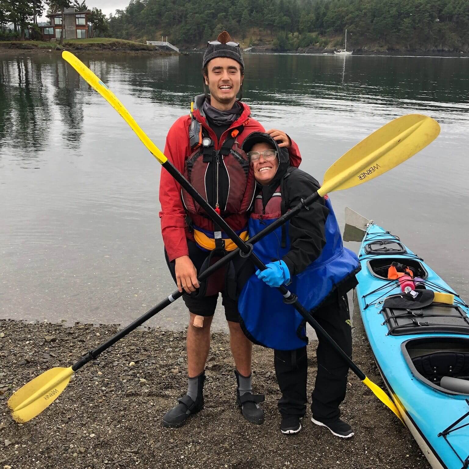 Two people in kayaking gear standing on a rocky shore smiling and holding yellow paddles with a blue kayak and calm water in the background