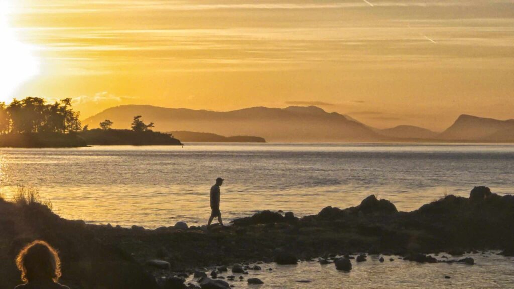 Adventure-vacation-gift-card | Sea Quest Kayak Tours A person walking along a rocky shore during sunset with a golden sky and silhouetted trees and mountains in the background