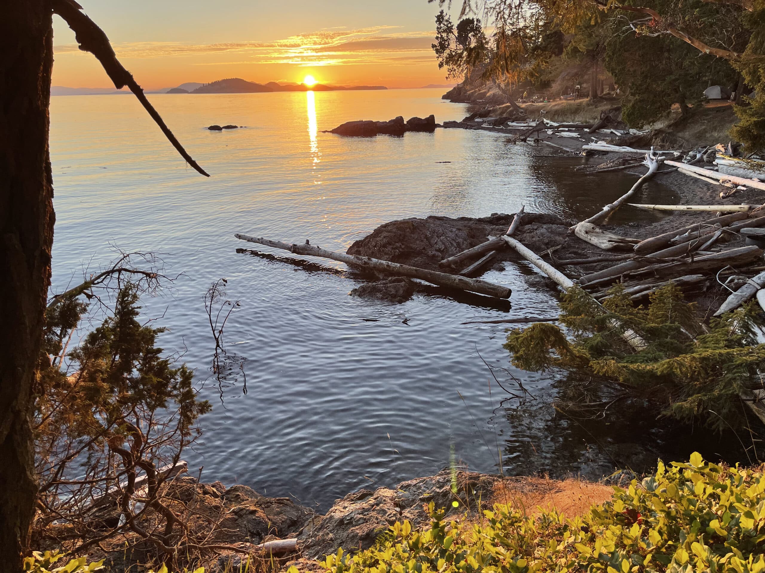 A scenic view of a sunset over the water with trees and rocky terrain in the foreground