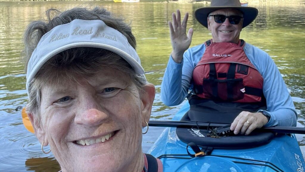 Close up selfie of a smiling woman wearing a visor with Hilton Head Island written on it with a man in the background wearing a hat sunglasses and a red life jacket sitting in a blue kayak and waving