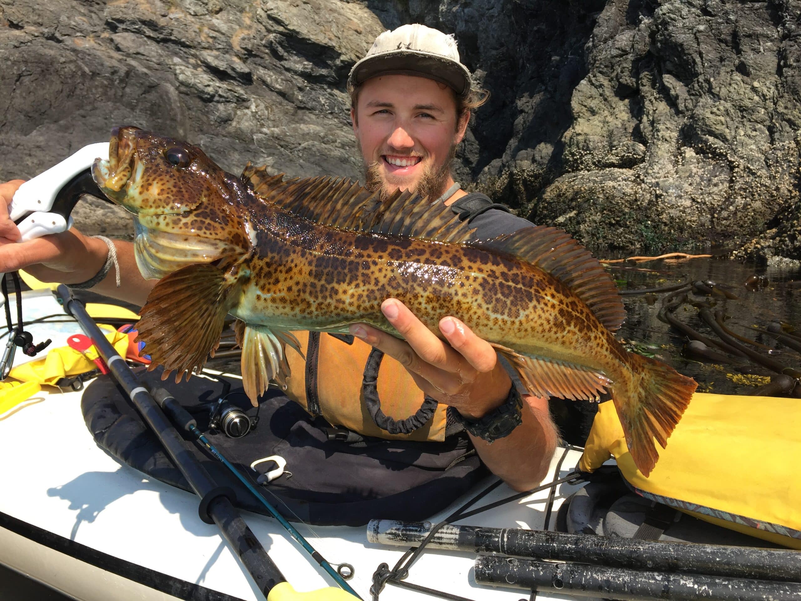 Man in a kayak holding up a large spotted fish he caught with rocky cliffs in the background and fishing gear visible in the kayak
