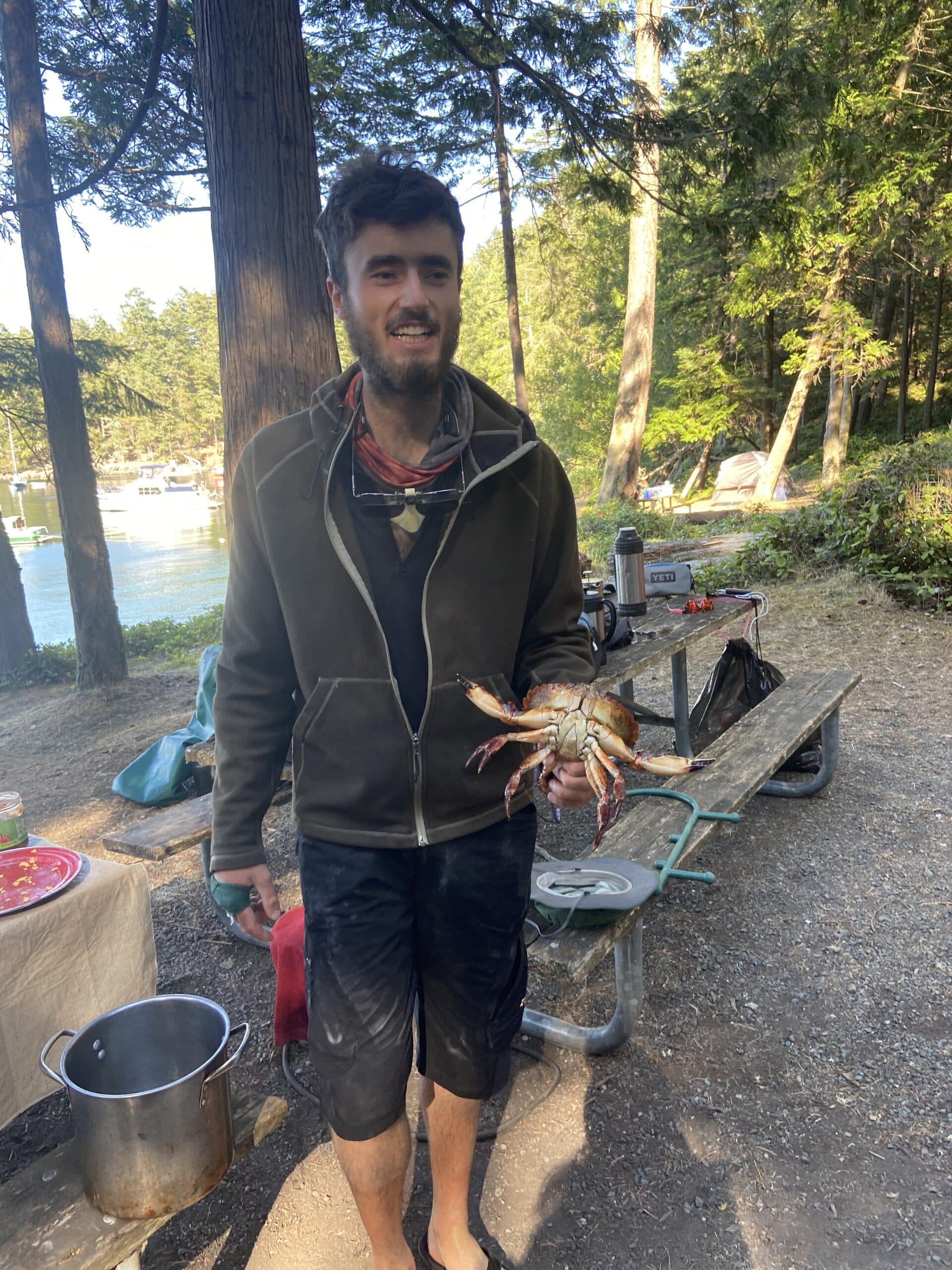 best-place-to-kayak-with-orcas | Sea Quest Kayak Tours A young man at a campsite holds a large crab, standing near a picnic table with cooking equipment. Tall trees and a glimpse of water with boats are in the background.