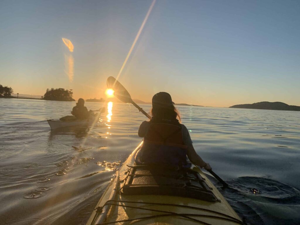 Two kayakers paddling on calm water at sunset with the sun low on the horizon and a golden glow reflecting on the water surrounded by distant islands and a clear sky
