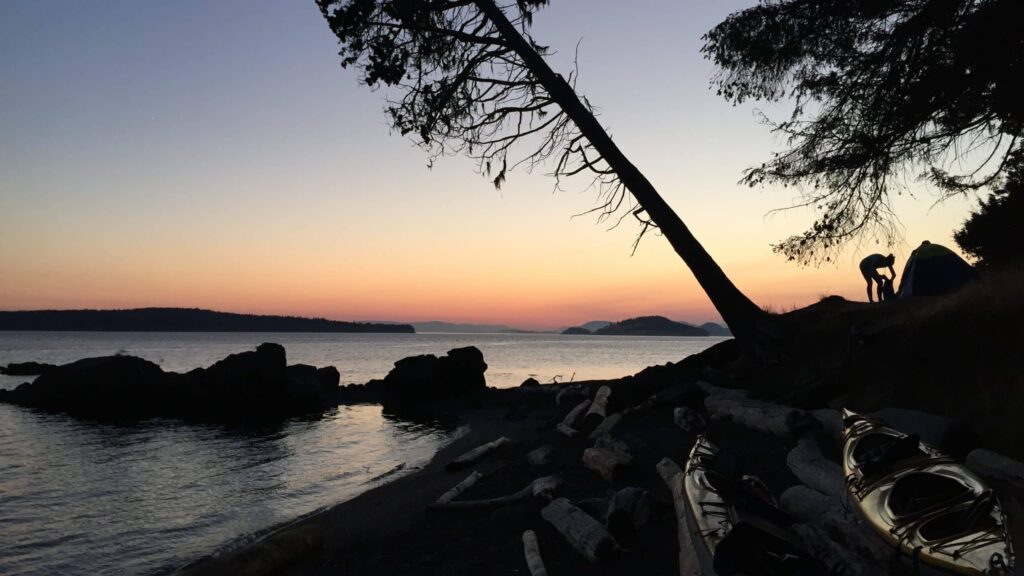 experienced-kayak-tour-company-san-juan-islands | Sea Quest Kayak Tours Scenic view of a shoreline at sunset with a silhouetted figure setting up a tent leaning trees kayaks on the ground and a calm body of water with distant islands under a colorful sky