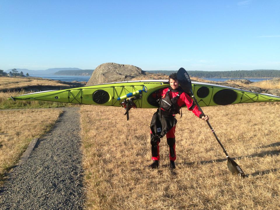 Person in red kayaking gear carrying a green kayak on their shoulders and holding a paddle standing on a grassy path with a rocky coastline and distant hills in the background under a clear blue sky