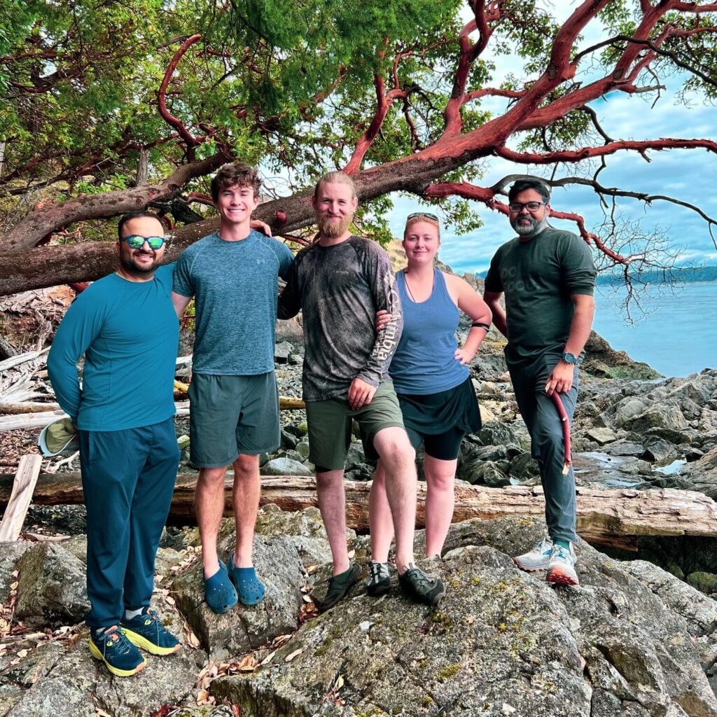 friday-harbor-things-to-do-hiking | Sea Quest Kayak Tours Group of five people standing on rocky terrain near a tree with red bark, posing for a photo with the ocean and a cloudy sky in the background.
