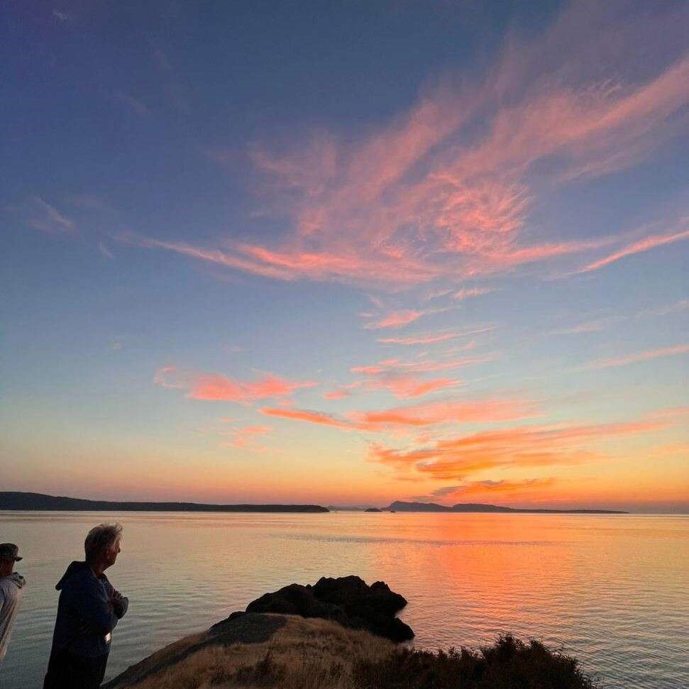 friday-harbor-things-to-do-lime-kiln-state-park | Sea Quest Kayak Tours Two people standing on a grassy cliff overlooking a calm body of water at sunset, with a colorful sky filled with pink and orange clouds reflecting on the water's surface.