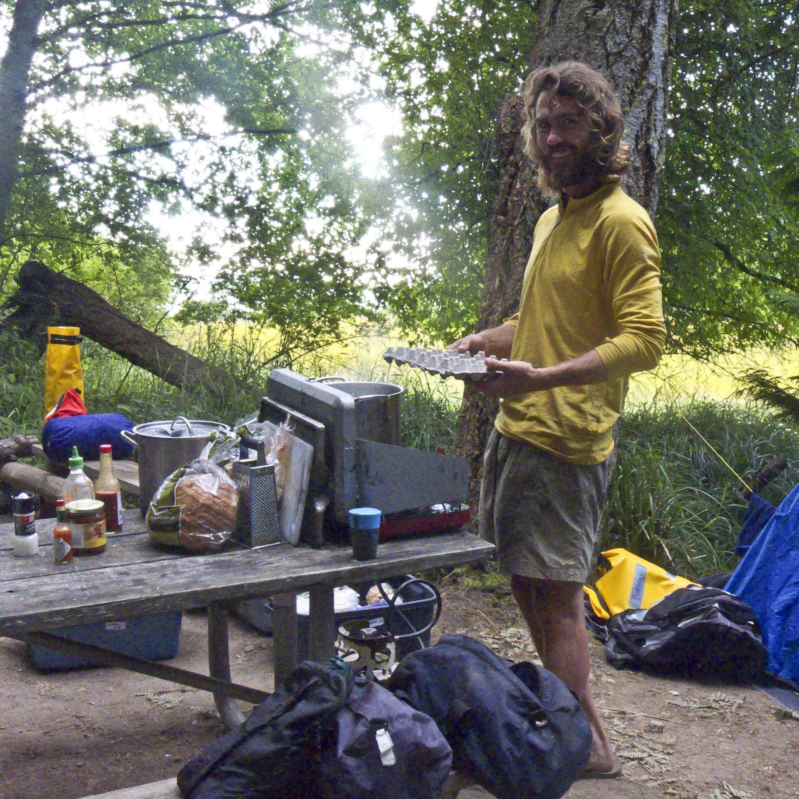 Man wearing a yellow long sleeve shirt standing at a picnic table in a wooded campsite preparing food with various supplies and cooking equipment on the table