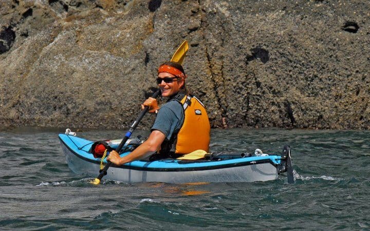 kayak-job | Sea Quest Kayak Tours A person in a kayak wearing a life jacket and a headband smiling and paddling near a rocky shoreline
