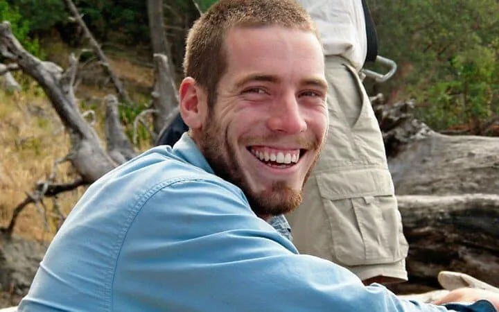 A smiling person wearing a light blue shirt seated outdoors with a backpack and trees in the background