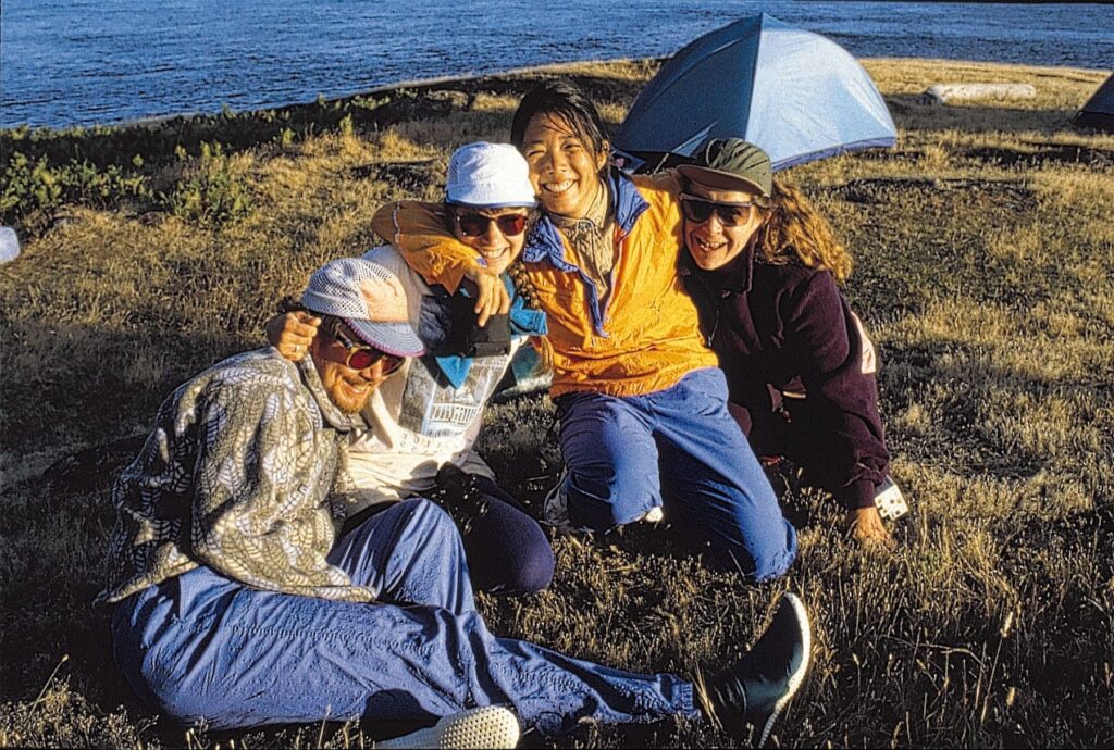 Four friends in outdoor gear sitting and laughing together on grassy terrain near the water with a blue tent in the background