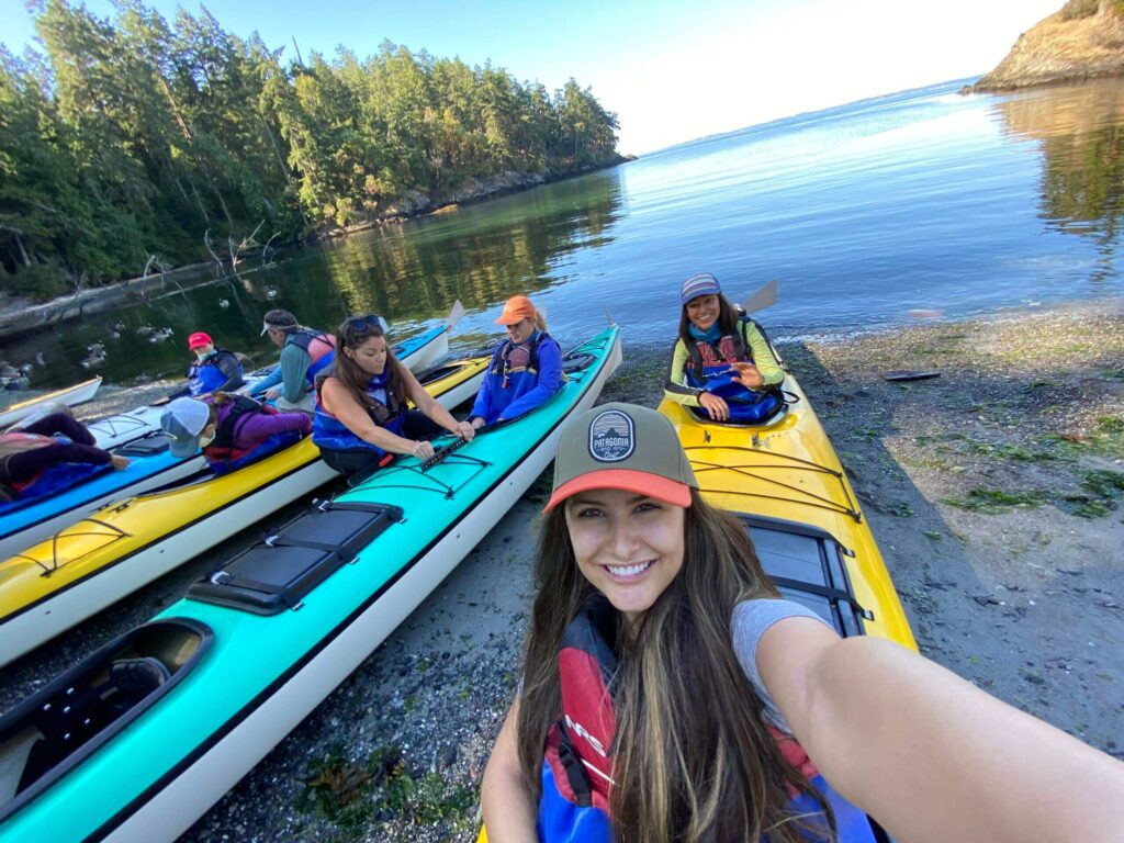 kayak-tours-excellent-reputation-san-juan-islands | Sea Quest Kayak Tours Group of people in colorful kayaks preparing for a kayaking trip with a woman in the foreground taking a selfie and smiling surrounded by a calm water inlet and dense forested area