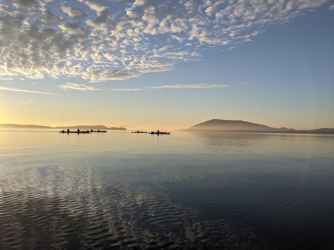kayak-vacation-san-juan-island-washington | Sea Quest Kayak Tours A serene view of kayakers paddling on calm water at sunset with a mountain and scattered clouds in the background