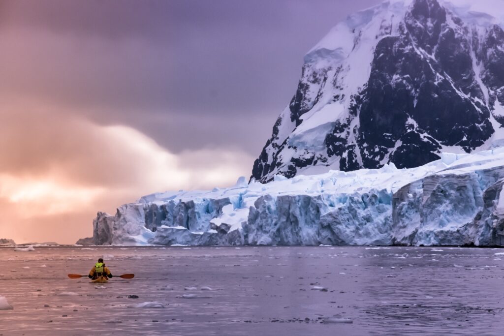 A person kayaking towards a massive snow-covered glacier and mountain under a dramatic sky with hues of purple and orange.