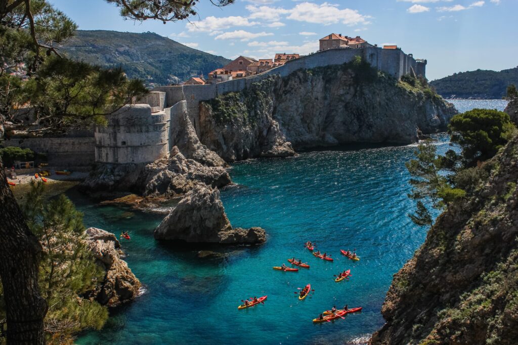 View of kayakers paddling in the clear, turquoise waters near the rocky coastline of a historic town with stone fortifications and terracotta-roofed buildings, surrounded by lush greenery and mountains under a blue sky with scattered clouds.