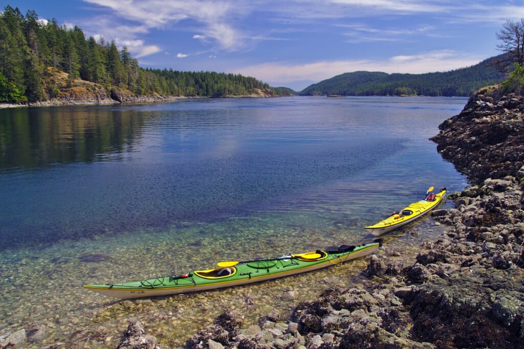 Two kayaks, one green and one yellow, rest on a rocky shore next to clear, calm water, surrounded by forested hills under a bright blue sky with wispy clouds.