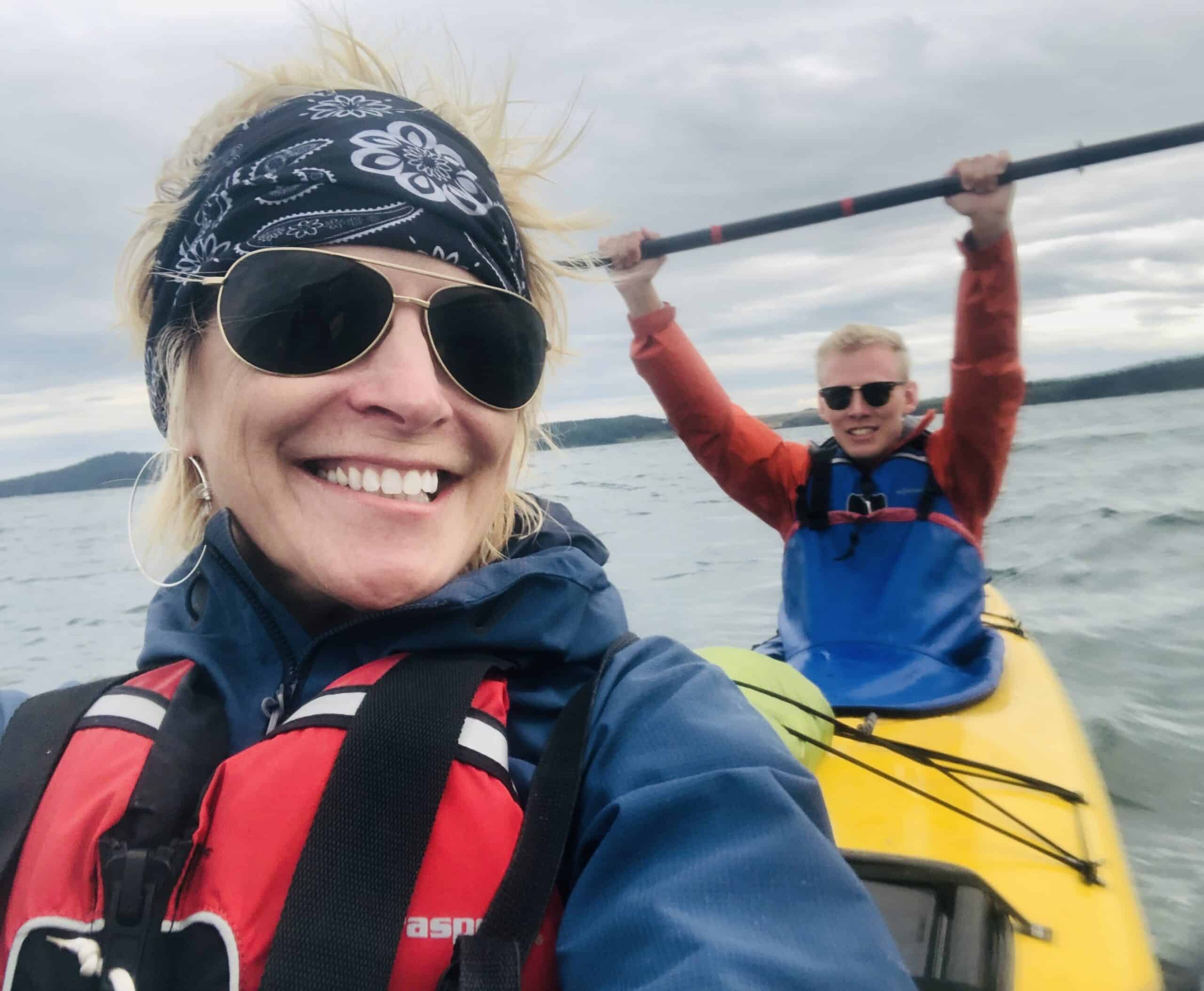 A woman smiles in the foreground wearing a red life jacket and sunglasses Behind her a man raises his paddle in a yellow kayak