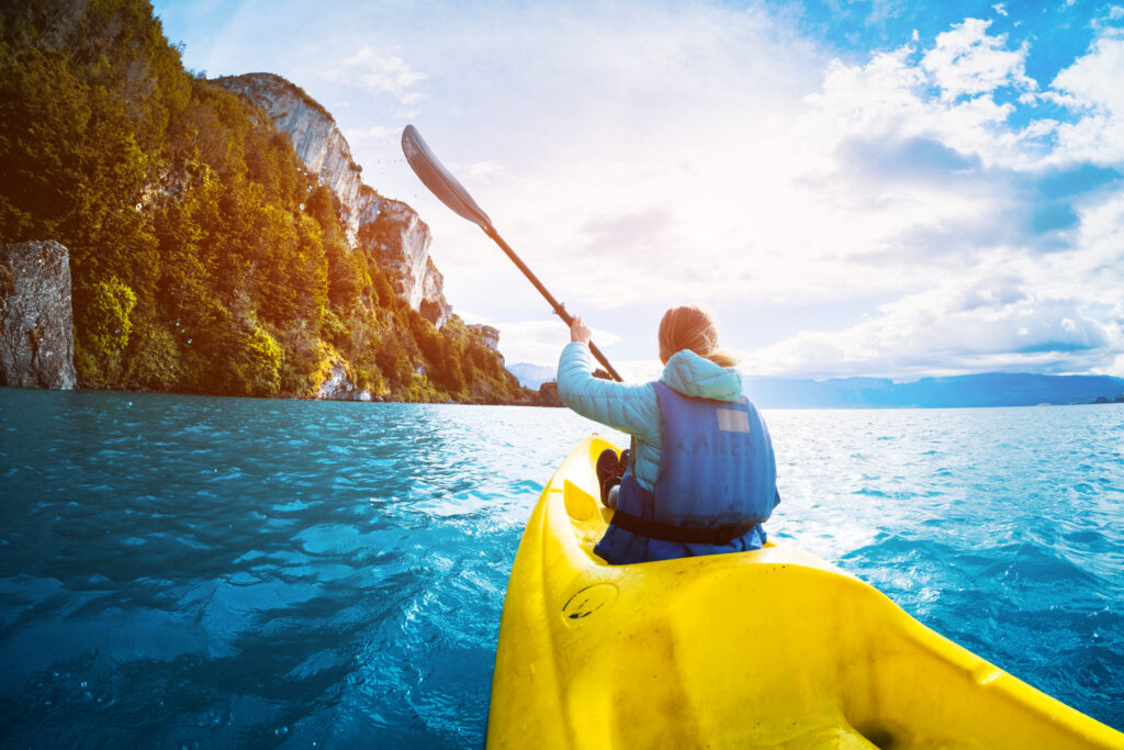 Person in a blue life jacket paddling a yellow kayak on a body of water, with green, rocky cliffs on the left and a bright sun shining through a partly cloudy sky.