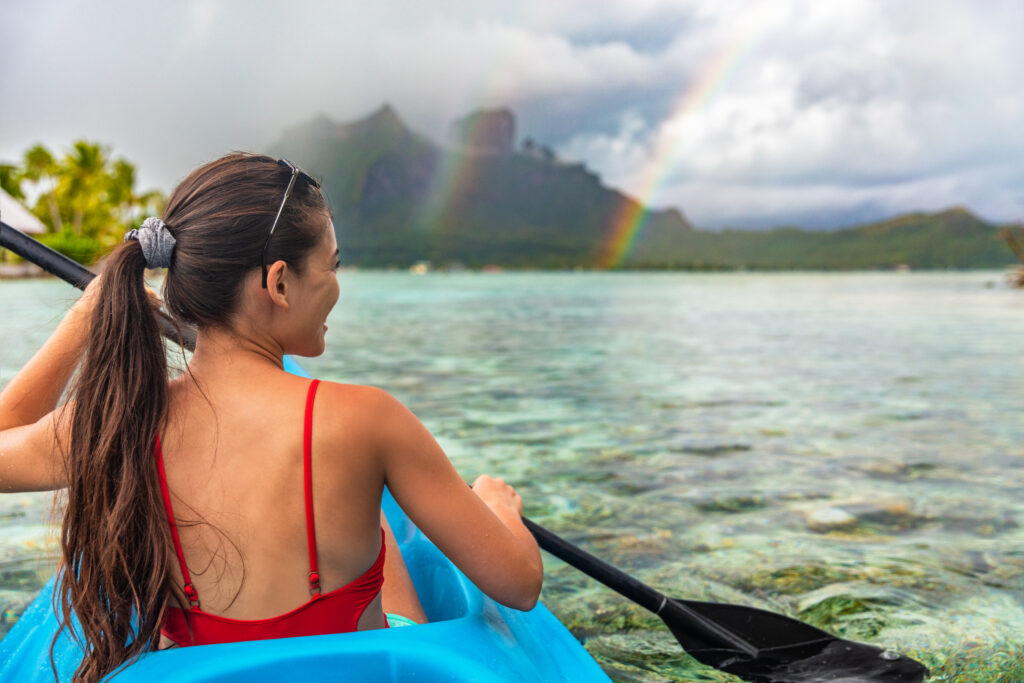 A woman in a red swimsuit paddling a blue kayak on clear, shallow water with a rainbow in the sky and misty mountains in the background.