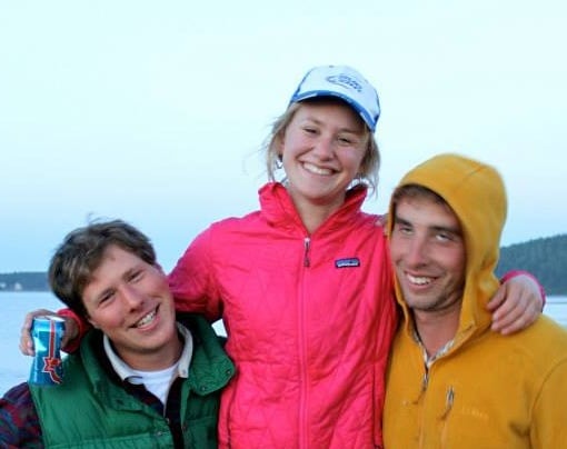 Three friends smiling with one person in a pink jacket and cap standing in the middle and two people in green and yellow jackets on either side
