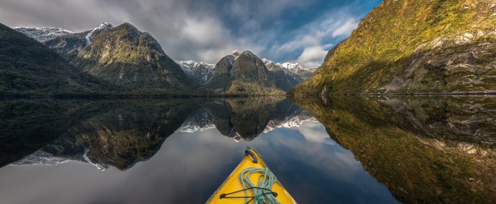 Panoramic,View,Of,Doubtful,Sound,,New,Zealand,,Seen,Over,The | Sea Quest Kayak Tours View from a yellow kayak on calm water, surrounded by tall, green mountains with snow-capped peaks, reflecting perfectly in the clear, still water under a partly cloudy sky.