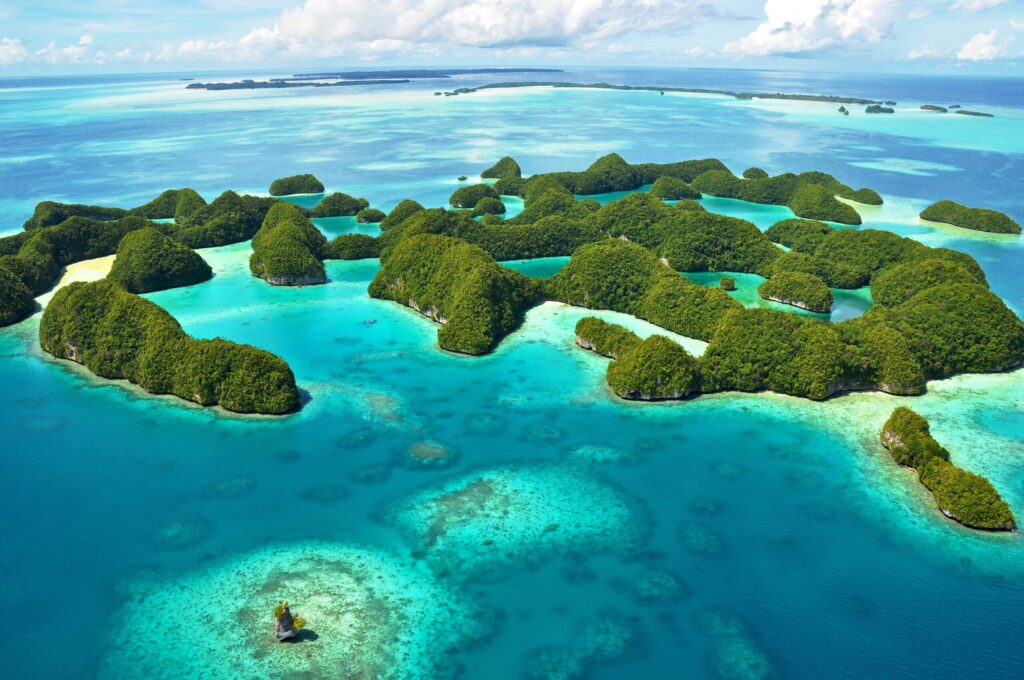 Aerial view of a group of lush, green islands surrounded by turquoise waters and coral reefs, with a larger expanse of blue ocean and distant islands under a partly cloudy sky.
