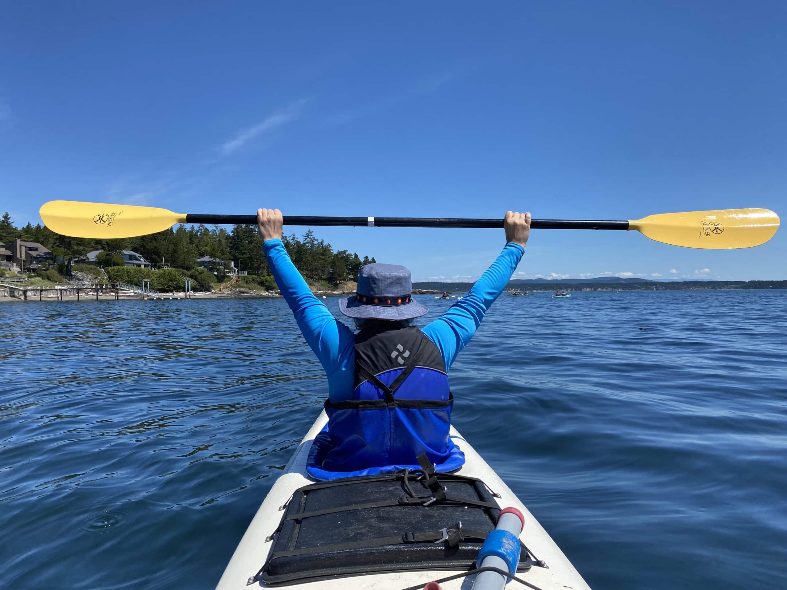 lightweight-kayak-paddle | Sea Quest Kayak Tours View from behind of a kayaker raising a yellow paddle triumphantly over their head while paddling on a calm blue sea. The kayaker is wearing a blue long-sleeve shirt, a life jacket, and a sun hat. A shoreline with houses and trees is visible in the distance under a clear blue sky.
