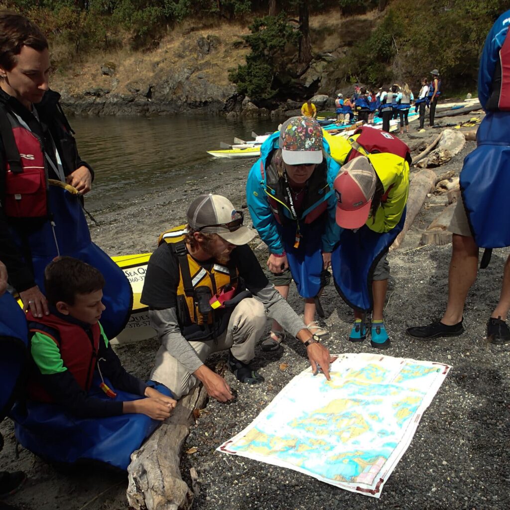 OLYMPUS DIGITAL CAMERA | Sea Quest Kayak Tours A group of people in outdoor gear gathered around a map on a rocky shore, planning their route.