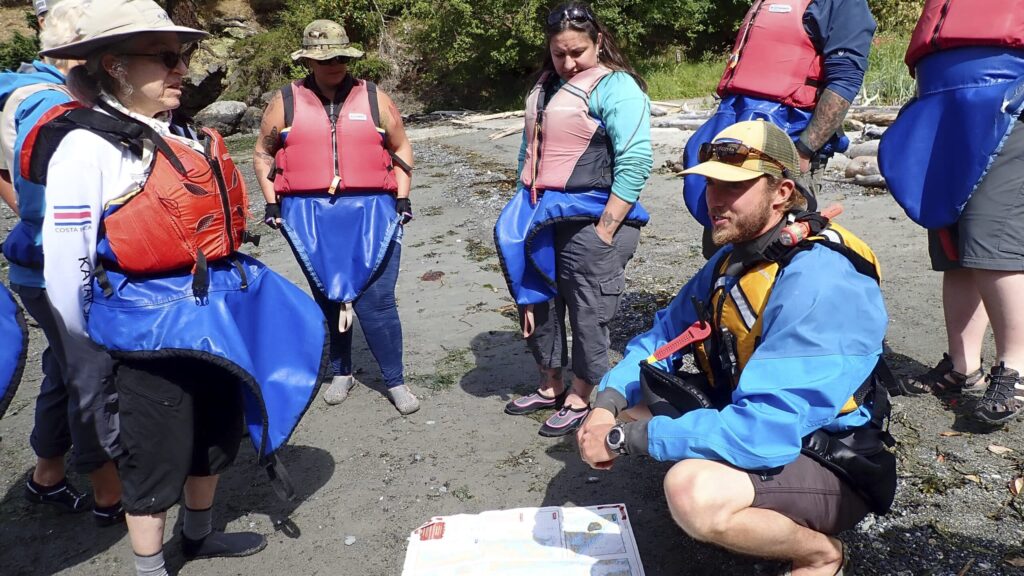 Group of people wearing life jackets and kayaking gear gathered on a beach with one person kneeling and explaining something using a map on the ground