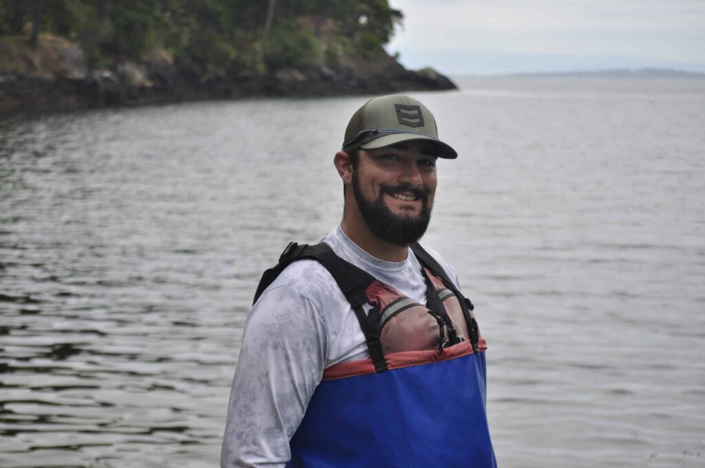 sea-kayaking-equipment-happy-customer | Sea Quest Kayak Tours Man smiling and standing near the water, wearing a green cap, a gray shirt, and a red and blue life jacket, with a rocky shoreline and trees in the background.
