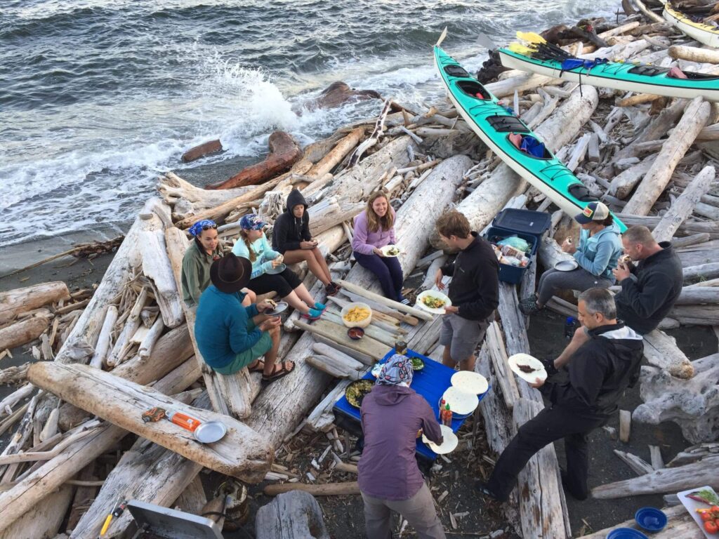 Group of people having a meal on a beach with driftwood logs and kayaks in the background sitting and standing around a makeshift table setup near the shoreline with waves crashing nearby