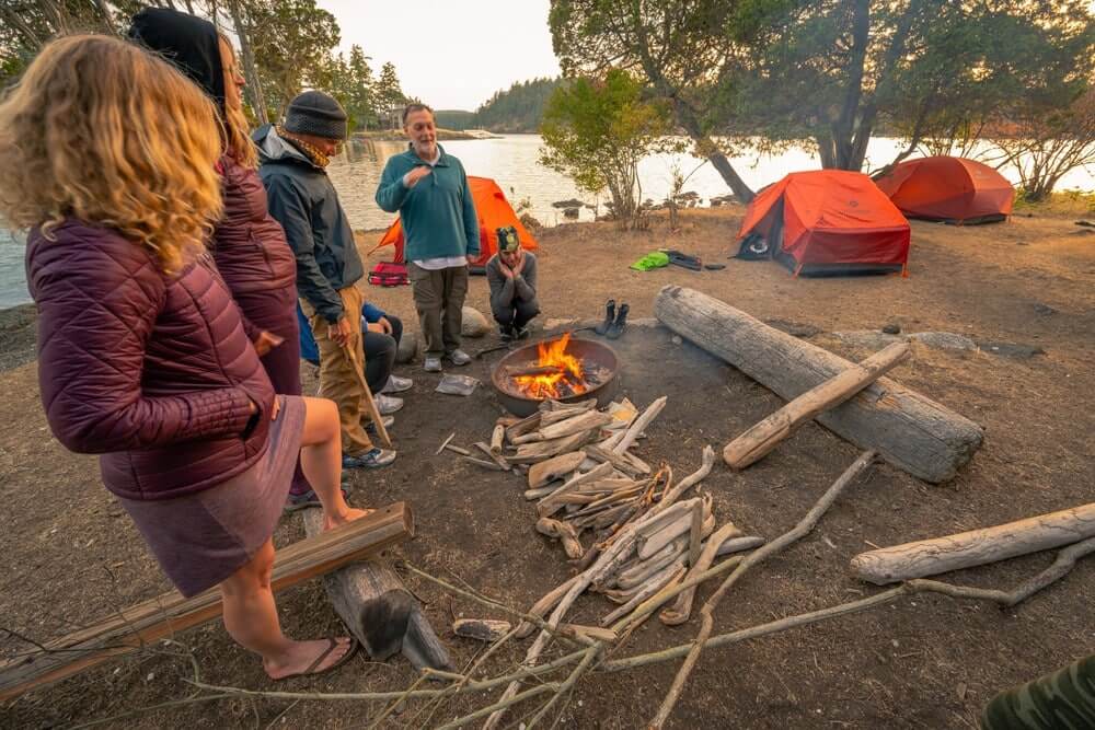 spacious-lighweight-tent-for-kayaking | Sea Quest Kayak Tours Campers gathered around a campfire near a body of water, with orange tents in the background.