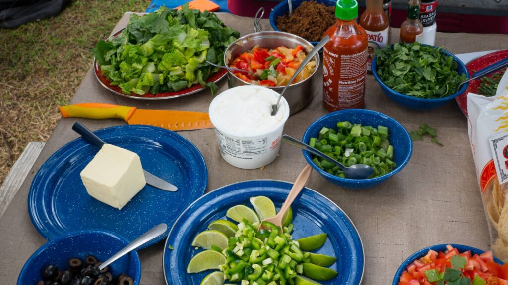 Close up of a picnic table with various ingredients and toppings for a meal including lettuce chopped bell peppers lime wedges green onions cilantro sour cream cheese black olives and hot sauces