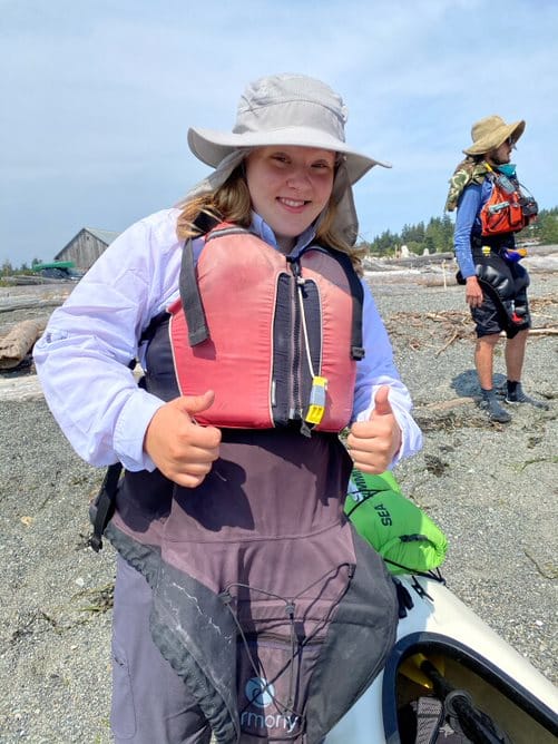 swimming-with-orcas | Sea Quest Kayak Tours A person wearing a red life jacket and sun hat gives a thumbs up while standing on a beach next to a kayak. Another person in outdoor gear is in the background.