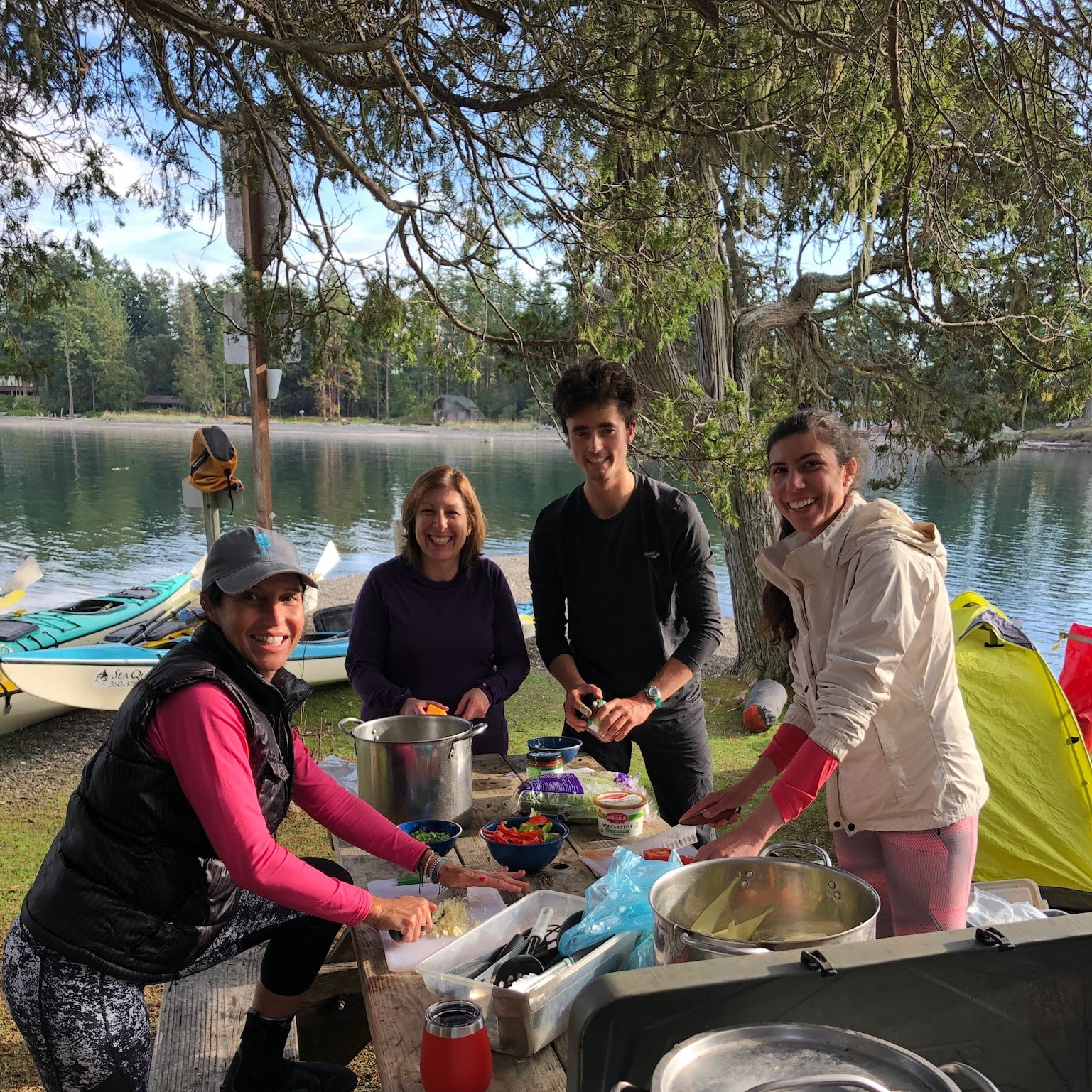 A group of friends smiling and sitting together at a campsite by the water with kayaks and gear visible behind them