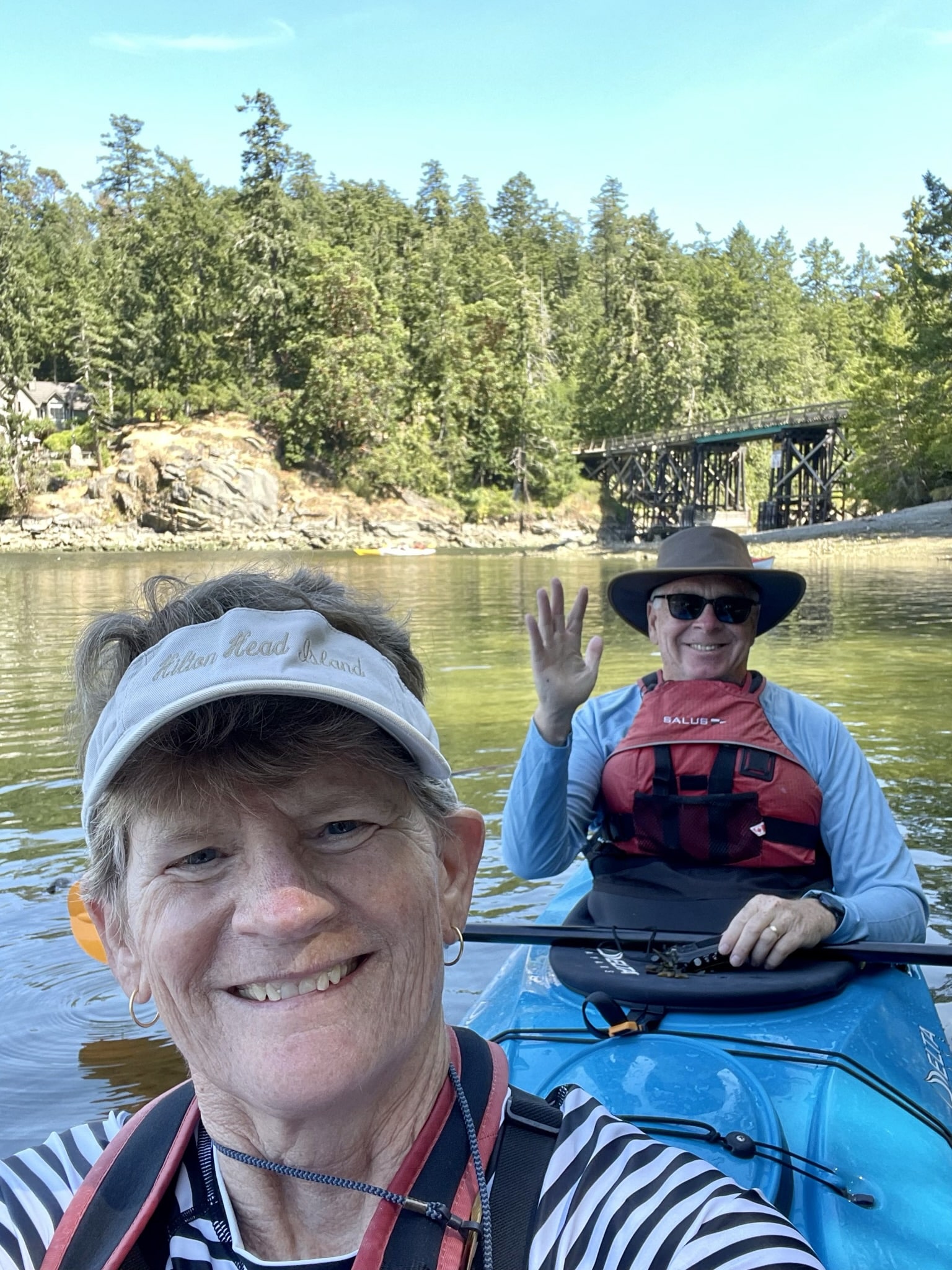 A person smiling and waving from a kayak on a calm river with trees and a bridge in the background