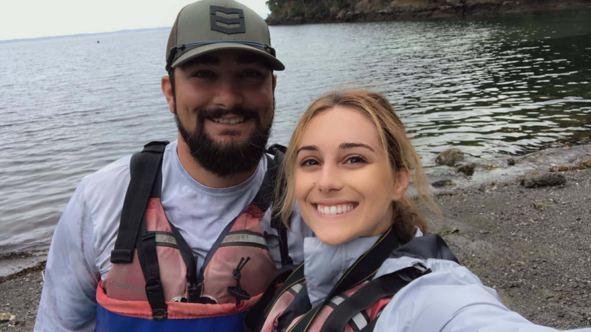A smiling couple in kayaking gear stands by the water