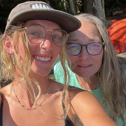 Two women smiling for a selfie the younger woman wearing glasses and a cap and the older woman wearing glasses and a light green shirt with trees and outdoor gear in the background