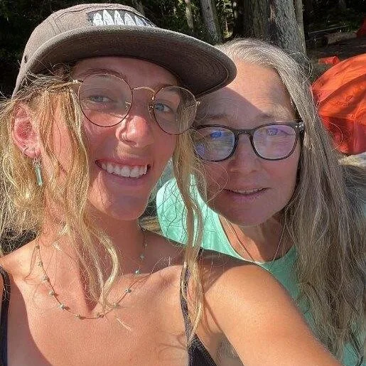 Two women smiling for a selfie the younger woman wearing glasses and a cap and the older woman wearing glasses and a light green shirt with trees and outdoor gear in the background