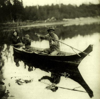 A historical black and white photo of a man a woman and a child fishing from a traditional wooden canoe on a calm river with a forested background