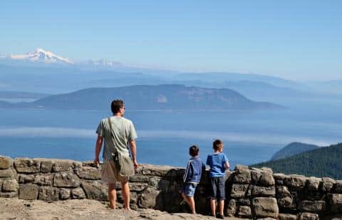 Mt-Constitution-Orcas-Island | Sea Quest Kayak Tours A man and two children stand at a stone railing looking out over a panoramic view of mountains islands and water under a clear blue sky