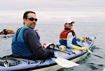 Two people sit in a tandem kayak on calm water They both wear life jackets and look back at the camera with the man in front wearing sunglasses and the woman behind him smiling