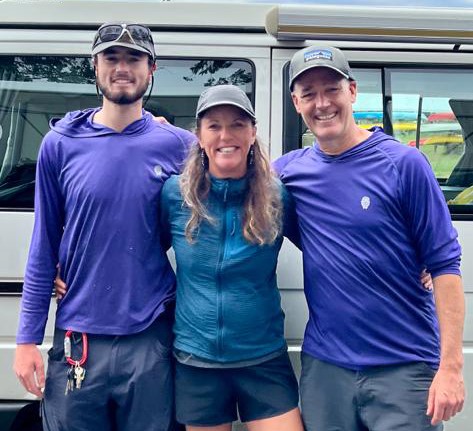 Three people standing in front of a vehicle smiling with their arms around each other the two men are wearing purple shirts and the woman in the middle is wearing a blue jacket and a cap