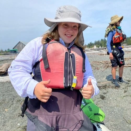 A young girl wearing a life jacket and hat, smiling at the camera, with another person in the background on a rocky beach.