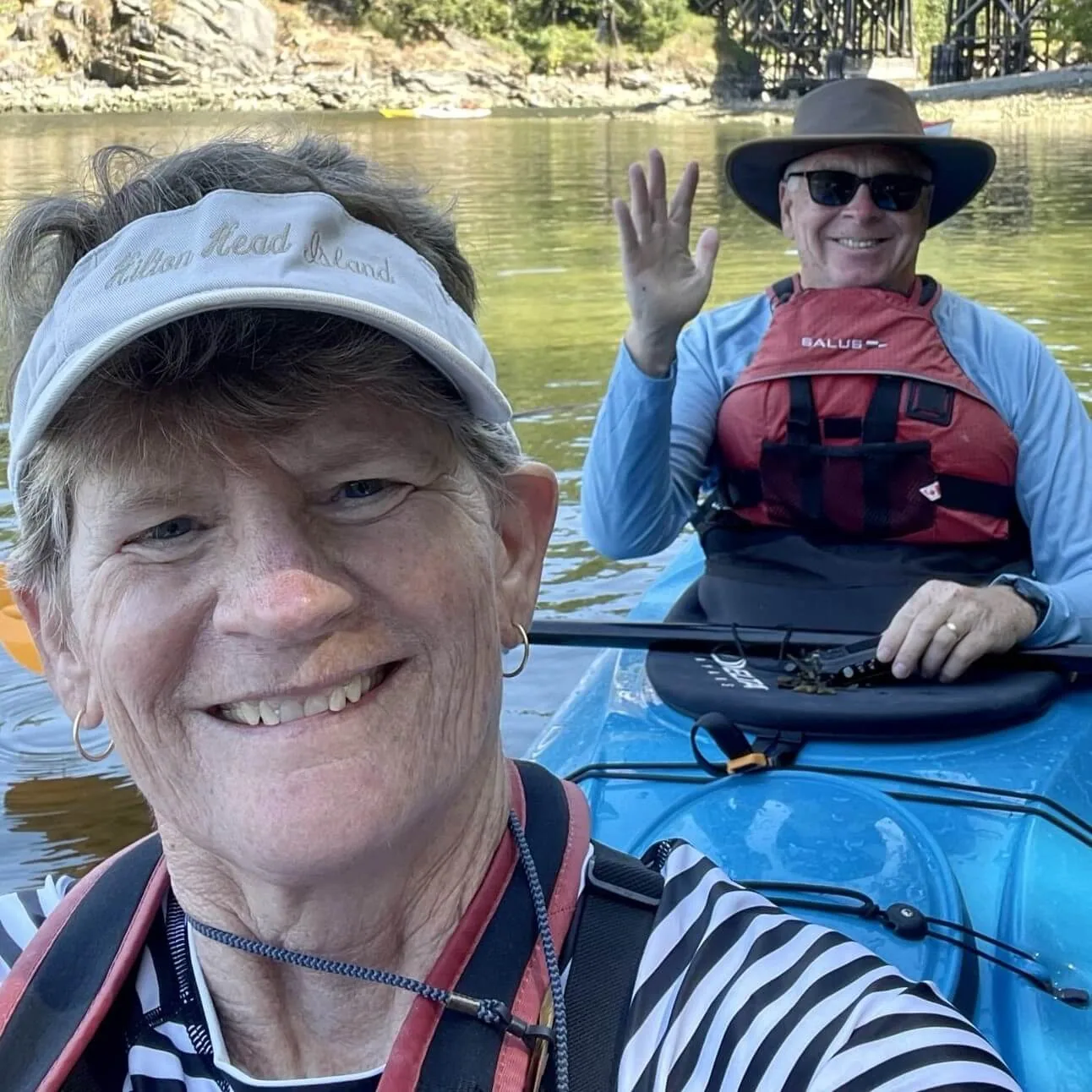 A woman and a man in a kayak on a calm river, with the man waving and both smiling at the camera.