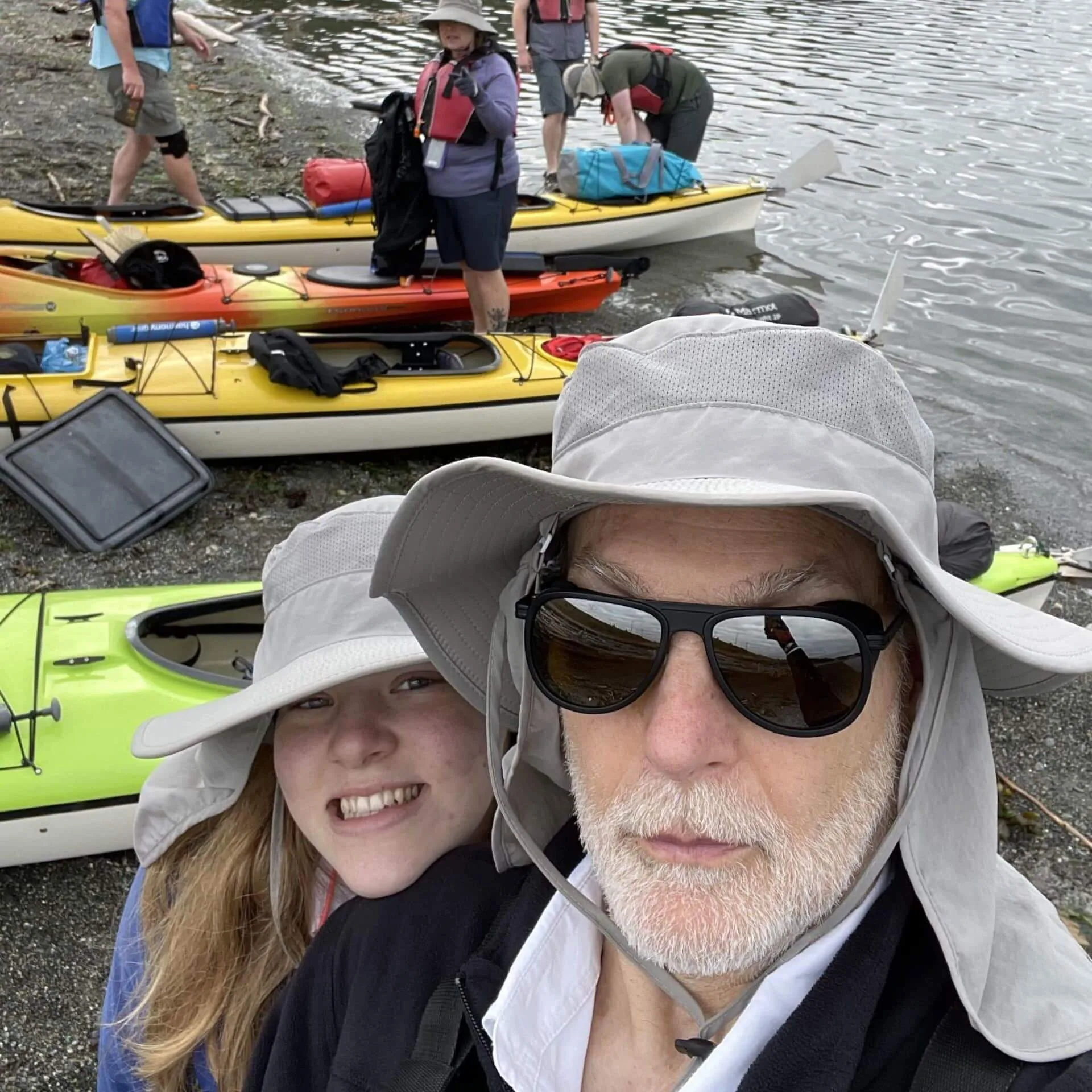 A man and a young girl smiling for a selfie in front of a group of kayaks on the shore.