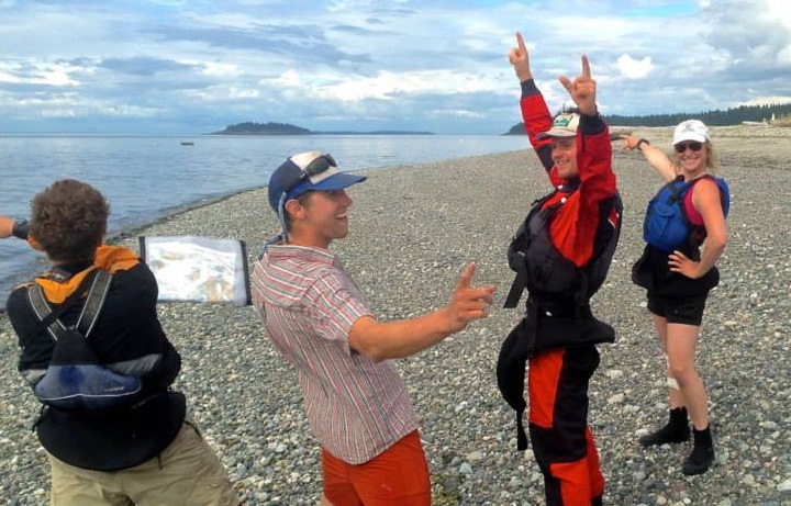 Four people standing on a rocky beach two in the foreground with one holding a map and two in the background one with arms raised in excitement
