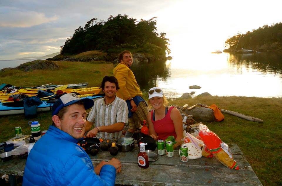 Four people sitting at a picnic table by the water with kayaks and gear in the background enjoying a meal outdoors at sunset