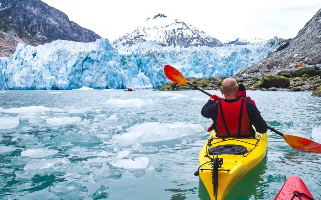 Person in a yellow kayak paddling towards a massive glacier, surrounded by floating ice chunks and towering snow-capped mountains.